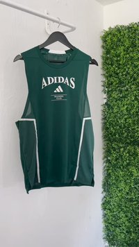 Zelena bezrukavna atletska majica s bijelim Adidas logotipom i natpisom, mrežastim bočnim panelima i kontrastnim bijelim obrubom na džepovima.