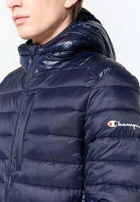 Námornícka puffa bunda s prešívaným dizajnom, kapucňou a bočným zipsom. Na ľavom rukáve je logo Champion. Ľahký, lesklý materiál.