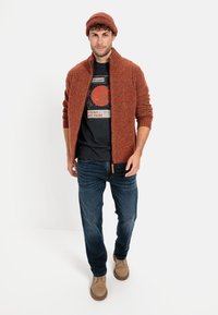 Rostfarbener Strickcardigan mit hohem Kragen, marineblaues Grafik-T-Shirt, blaue Jeans und sandfarbene Stiefel. Das Ensemble wird durch eine passende Mütze ergänzt.