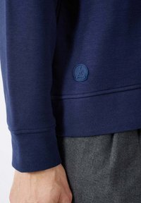 Sudadera azul navy con tejido texturizado, puños acanalados y un logotipo redondo en relieve. Combinada con pantalones grises texturizados.