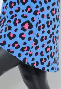 Blaues Oberteil mit schwarzen und pinken Leopardenpunkten, mit einem asymmetrischen Saum und Seitennähten. Kombiniert mit glänzenden schwarzen Leggings.