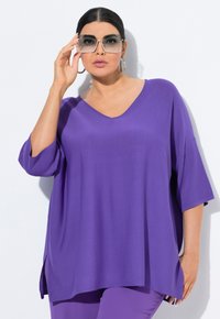 Lila übergroße Bluse mit V-Ausschnitt, kurzen Ärmeln und seitlichen Schlitzen. Das Model trägt große Sonnenbrillen und Creolen. Glatte Stofftextur.