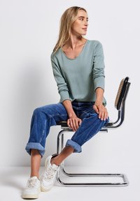 Lichtgroen langmouwshirt, losse blauwe jeans met opgerolde uiteinden, en witte sneakers, zittend op een chroom en geweven stoel tegen een witte achtergrond.