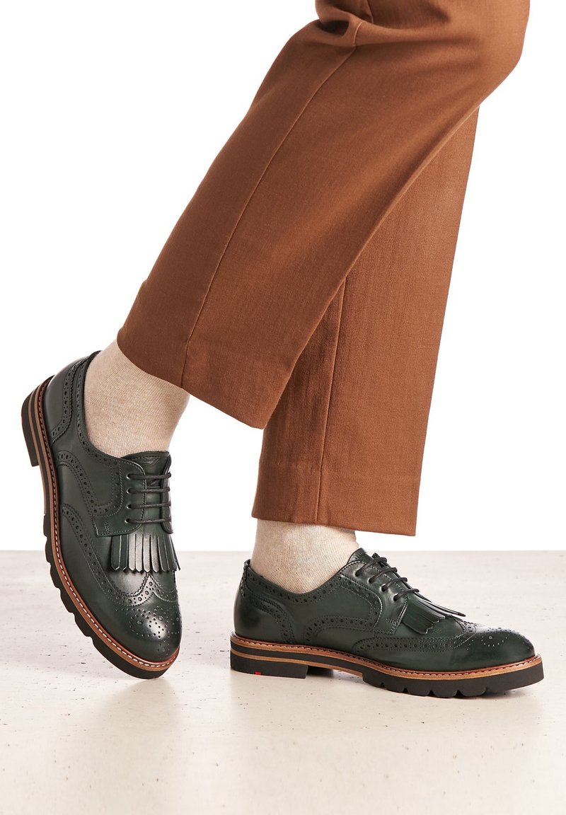 Brogues en cuir vert avec des détails perforés et des pompons, présentant un bout rond et une semelle en caoutchouc noire, associés à un pantalon marron raccourci.