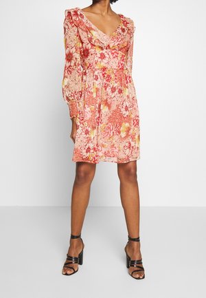 Femme portant une robe fleurie mi-longue à manches longues avec un col à volants et des sandales noires à talons hauts et brides sur un fond blanc.