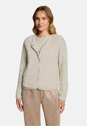 Frau mit schulterlangem blondem Haar, die eine hellbeige strukturierte Wollweste über einem langärmligen Stricktop und beigen Hosen trägt.