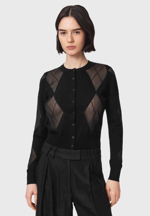 Vrouw met kort donker haar, die een zwarte, geknoopte cardigan draagt met een diamanten doorzichtig patroon en een zwarte broek met hoge taille.