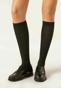 Czarne skarpetki sięgające do kolan mają teksturowany wzór z pionowymi liniami. W połączeniu z błyszczącymi czarnymi mokasynami. Cały design jest elegancki i dopasowany.