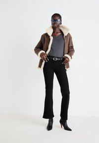 Jachetă din shearling maro cu căptușeală crem, bluză gri cu decolteu în V, blugi negri evazați și pantofi cu toc ascuțit negri. Model care poartă ochelari de soare.