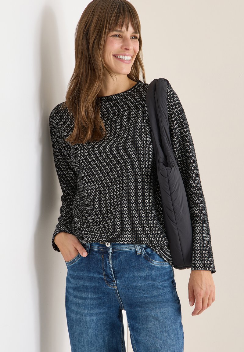 Top a maniche lunghe a fantasia nera e crema abbinato a jeans blu, con un leggero giubbino nero appoggiato su una spalla.