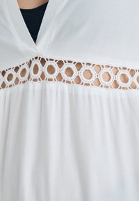 Top branco com decote em V e um painel de renda decorativo com recortes circulares. Tecido suave com uma textura leve e fluida.