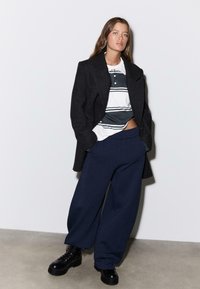 Crni vuneni kaput preko bijele i tamnoplave polo majice s prugama, uparen s velikim tamnoplavim trenirkama i masivnim crnim čizmama.