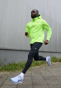 Giacca da corsa verde neon con cappuccio, leggings neri e sneaker bianche con dettagli blu. Il modello è in una posa da corsa su un sentiero.