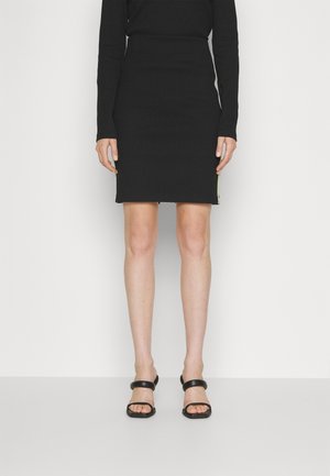 Personne portant une robe noire à manches longues et des chaussures à talons ouverts noires, debout devant un fond clair uni.