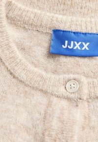 Beige stickad cardigan i nära vy, med rund halsringning, en liten knapp och en blå etikett med "JJXX" i vit text. Mjuk textur.