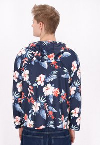 Heren bloemerige hooded jack in navy met witte en rode hibiscussen en groene bladeren; uitgerust met lange mouwen, lichtgewicht stof en een casual pasvorm.