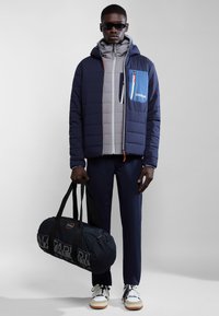 Giacca a vento navy con fodera interna grigia e tasca blu, maglia grigia, pantaloni scuri e borsa da palestra nera con logo. Sneakers bianche.