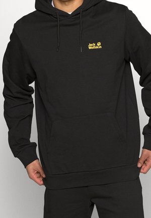 Person iført en sort Jack Wolfskin hættetrøje med frontlomme og gul logo på brystet, der holder hættetrøje-kanten med begge hænder.