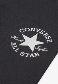 Svart stoff med en hvit sirkulær logo som har en stjerne og teksten: "CONVERSE Chuck Taylor ALL STAR." Teksten har en blandet skrifttype.