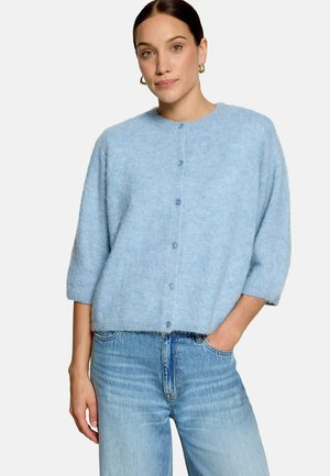 Femme portant un cardigan bleu clair en maille duveteuse boutonné avec des manches trois quarts et un jean bleu clair taille haute, debout avec une main dans la poche.