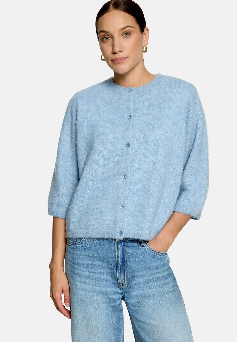 Femme portant un cardigan bleu clair en maille duveteuse boutonné avec des manches trois quarts et un jean bleu clair taille haute, debout avec une main dans la poche.