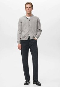 Cardigan gris hecho de material suave, con cierre de botones, cuello redondo, puños acanalados, combinado con jeans oscuros y zapatos negros.