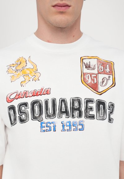 T-shirt blanc à manches courtes avec des graphiques colorés : un lion, "Canada," "DSQUARED2," et un emblème avec des chiffres. Matière en coton avec une texture douce.