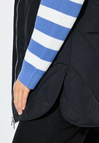 Chaleco acolchado negro con un acabado mate, que presenta un cierre de cremallera y un dobladillo con bordes redondeados, combinado con una manga de punto a rayas en azul y blanco.
