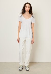 Bijela pamučna majica s V-izrezom i kratkim rukavima, u kombinaciji s svijetlim ravnim trapericama i sivim tenisicama s žutim naglascima.