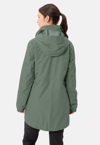 Lange, kapuzenjacke aus einem gedämpften grünen Stoff. Verfügt über elastische Ärmelbündchen, eine Länge, die die Hüften bedeckt, und ein Mesh-Futter, das an der Kapuze sichtbar ist.