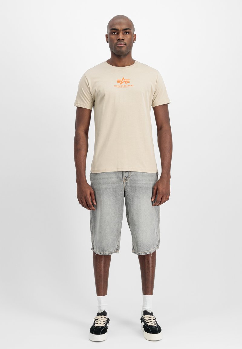 Camiseta de algodón beige con un logo naranja prominente en el pecho, combinada con pantalones cortos de mezclilla grises y zapatillas negras con detalles blancos.