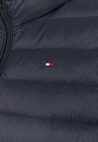 Ciemnogranatowa kurtka puchowa z pikowanymi panelami i małym haftowanym logo w kolorach czerwonym, białym i niebieskim. Gładki, lekki materiał.