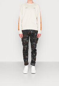 Sudadera gris claro con rayas naranjas en las mangas y logo. Pantalones cargo camuflados negros con múltiples bolsillos. Zapatillas blancas.