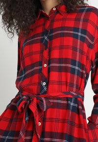 Abito camicia a quadretti rossi e blu con bottoni frontali, colletto e vita legata. Realizzato in un tessuto dalla consistenza morbida e dal design morbido.