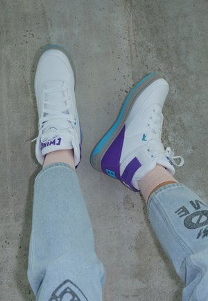 Osoba nosząca białe trampki z fioletowymi i niebieskimi akcentami oraz jasnoniebieskie dżinsy na szarej betonowej podłodze.