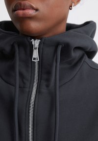 Svart zip-up-hoodie med hög krage, utrustad med en silverfärgad dragkedja och dragsnören. Materialet verkar mjukt med en slät yta.