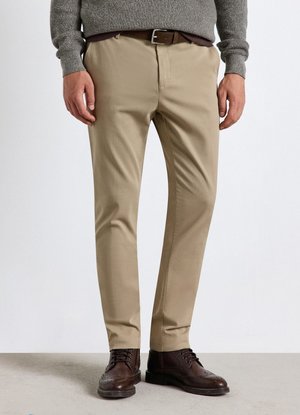Uomo che indossa pantaloni beige con cintura nera, maglione grigio e scarpe eleganti marrone scuro, in piedi su un pavimento chiaro davanti a uno sfondo semplice.
