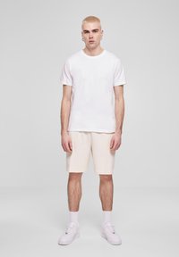 Homme blond debout, portant un t-shirt blanc, un short crème, des chaussettes blanches et des baskets blanches, devant un fond gris clair uni.