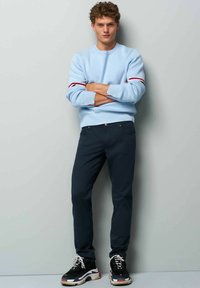 Lichtblauwe crewneck sweatshirt met rode en witte strepen, gecombineerd met donkerblauwe broek en zwarte sneakers met multikleurige zolen.