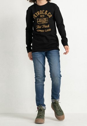 Osoba nosząca czarną koszulkę z długim rękawem z żółtym napisem, niebieskie dżinsy i zielone sznurowane buty, stojąca na jednolitym białym tle.