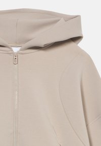 Μπεζ φούτερ με κουκούλα και μπροστινό φερμουάρ, από μαλακό ύφασμα και στρογγυλεμένους ώμους. Διαθέτει λεία υφή και μινιμαλιστικό σχέδιο.