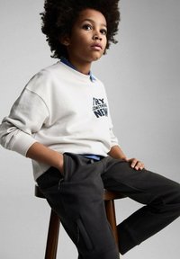 Vit sweatshirt med trycket "PROVA NÅGOT NYTT", i kombination med mörkgrå byxor med fickor. Barn med lockigt hår som sitter på en trähylla.
