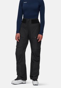 Zwarte geïsoleerde skibroek met een hoge taille, klittenbandaccenten, meerdere met rits afgesloten zakken en versterkte enkelpatches. Gevoerd met witte skischaatsen.