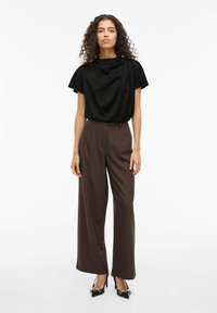 Top nero drappeggiato con maniche corte, abbinato a pantaloni larghi a vita alta marroni. La modella indossa décolleté nere a punta con dettagli dorati.