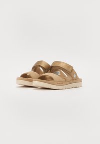 Beige sandaler med et strukturert stoffoverlegg, justerbare stropper, åpen tå og en formet gummisåle med et sik-sak mønster.