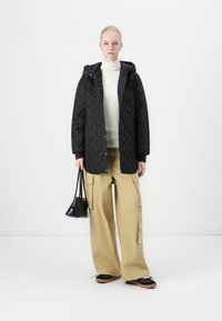 Chaqueta acolchada negra con capucha, suéter de cuello alto blanco debajo, pantalones cargo de pierna ancha beige y un bolso negro pequeño. Zapatos negros con suelas marrón.