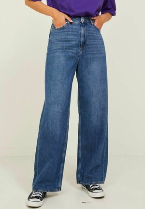 Jeans in denim blu a vita alta e gamba larga con design a cinque tasche e una leggera sfumatura, abbinati a sneakers in tela nera.
