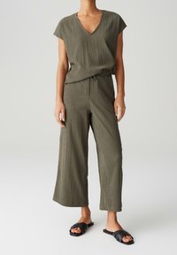 Olivově zelený overal s texturou krabicového charakteru, s výstřihem do V, širokými nohavicemi a elastickým pasem. V kombinaci s černými nazouvacími sandály.