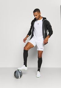 Zwarte zip-up jas, witte t-shirt, witte shorts, zwarte kniekousen en witte sneakers met groene accenten, staand boven een grijze voetbal.