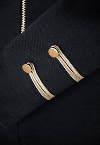 Czarna odzież z złotymi akcentami na rękawach, charakteryzująca się teksturowanym materiałem i dekoracyjnym sznurem przymocowanym do okrągłych guzików.